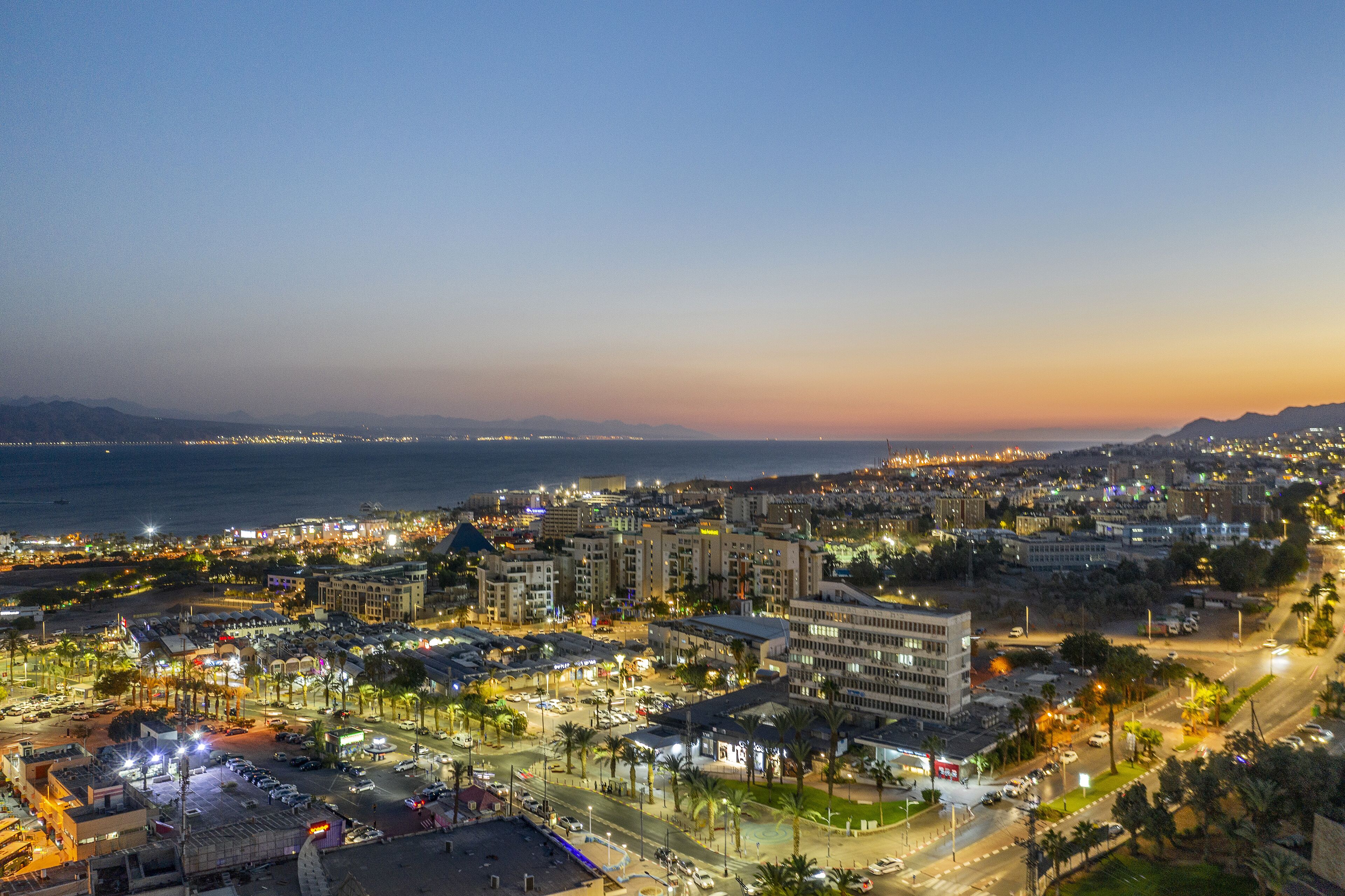 A cityscape at dusk with illuminated buildings, roads, and a coastline.