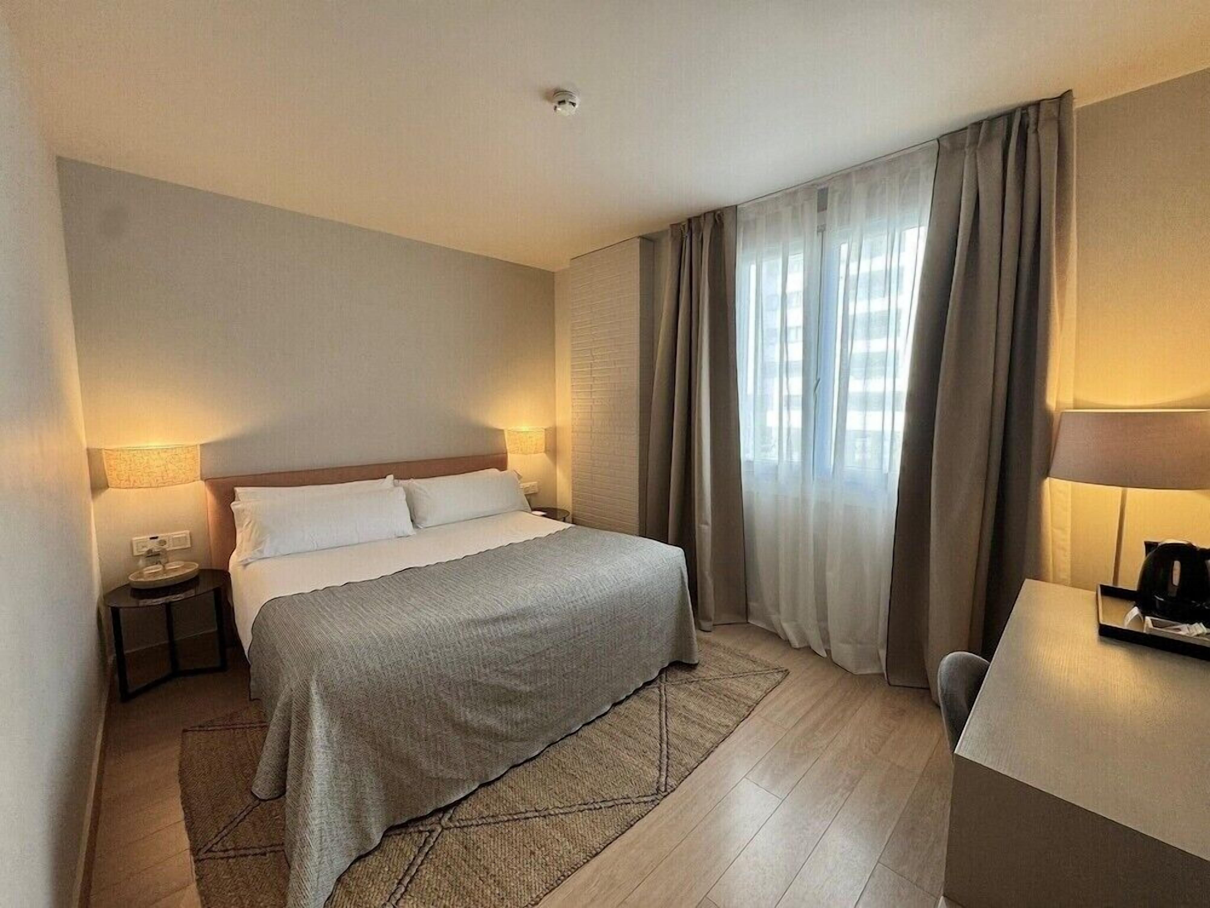 A hotel room with a bed, bedside tables, a desk, and a window with curtains.