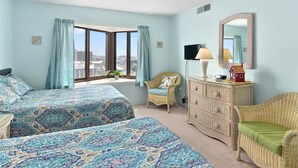 A bedroom with two beds, a dresser, a chair, a lamp, and a large window offering a city view. - Sweeping ocean and bayviews, large outdoor pool,2 Br/2 Ba. (Ocean City)