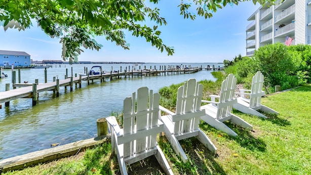 Blick auf das Wasser mit einer anlegenden Mole, einer Reihe Adirondack-Stühle und einem Gebäude mit Balkonen.