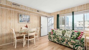 A room with a wooden floor, a white sofa with a tropical print, a dining table with chairs, and a door with a number.