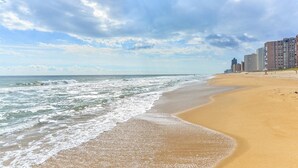 A beach with a sandy shore and waves, a clear sky, and buildings in the background.