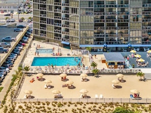 A hotel with a swimming pool, beach area with sun loungers and umbrellas, and a parking lot.