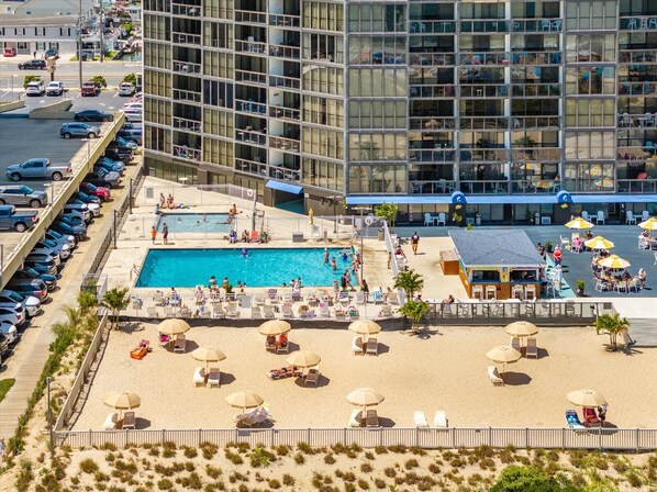 A hotel with a swimming pool, beach area with sun loungers and umbrellas, and a parking lot.