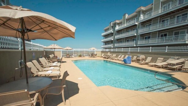 A pool area with lounge chairs and umbrellas, surrounded by a multi-story building with balconies. - Pretty 2 br/ 2 bath with tranquil bayviews, easy walking to the beach and outdoor pool. (Ocean City)