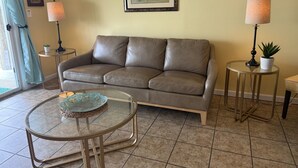 A living room with a brown leather sofa, a glass coffee table, two side tables with lamps, a framed painting, and a potted plant.