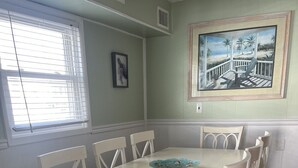 Een eetkamer met een witte tafel en stoelen, een lijst met een afbeelding aan de muur, en een raam met jaloezieën.