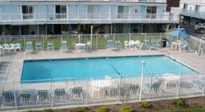 Una piscina rettangolare con un'area recintata, circondata da sedie e tavoli bianchi, e un edificio con balconi sullo sfondo.