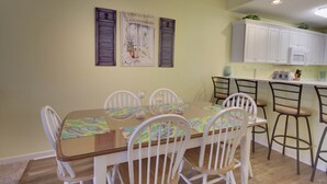 A dining area with a glass-top table, white chairs, and a kitchen area with white cabinets and a microwave. - Beautiful condo, located 61st St ocean block. 3 br, with outdoor pool. (Ocean City)