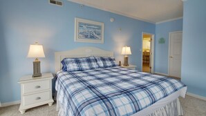A bedroom with a blue and white checkered bedspread, white nightstands with lamps, and a white door. - Beautiful condo, located 61st St ocean block. 3 br, with outdoor pool. (Ocean City)