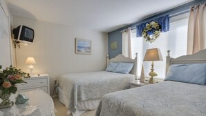 A bedroom with two beds, a television on a shelf, a bedside table with a lamp, a framed picture on the wall, and a window with curtains.