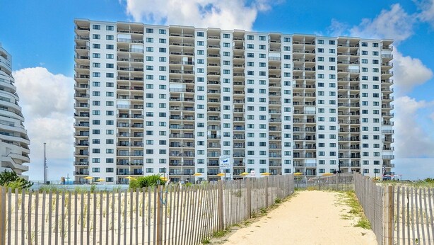 A high-rise hotel with multiple balconies, a sandy pathway leading to the entrance, and a clear view of the sea.