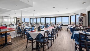 A spacious dining area with tables and chairs, large windows offering a view of the sea, and a wooden floor.