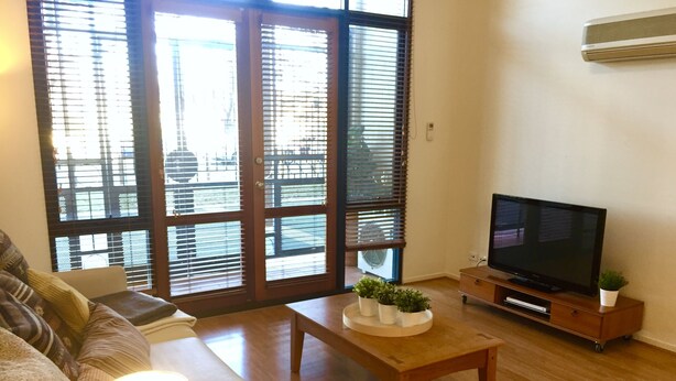 A living room with a sofa, a coffee table with plants, a television on a wooden stand, and large windows with blinds. - Central Loft Apartment Free Parking (Canberra)