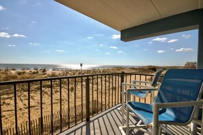A balcony with blue chairs overlooking a beach with sand dunes and a fence.