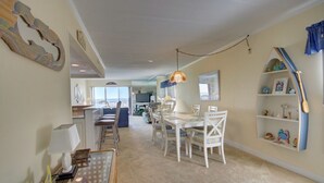 A spacious living area with a dining table and chairs, a bookshelf, and a large window providing a view of the outdoors. - Sand Trap 102 - 2br/2ba oceanfront in area of low beach congestion. (Ocean City)