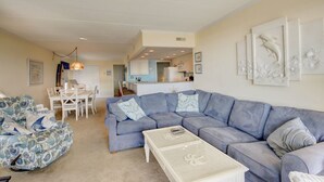 A living room with a blue sofa, a patterned armchair, and a coffee table. There is a dining area with a table and chairs. - Sand Trap 102 - 2br/2ba oceanfront in area of low beach congestion. (Ocean City)