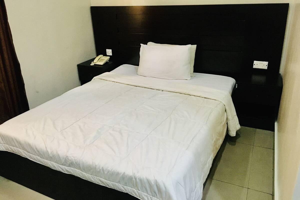 Cama bem arrumada com roupa de cama branca, cabeceira preta e mesa de cabeceira com telefone branco.