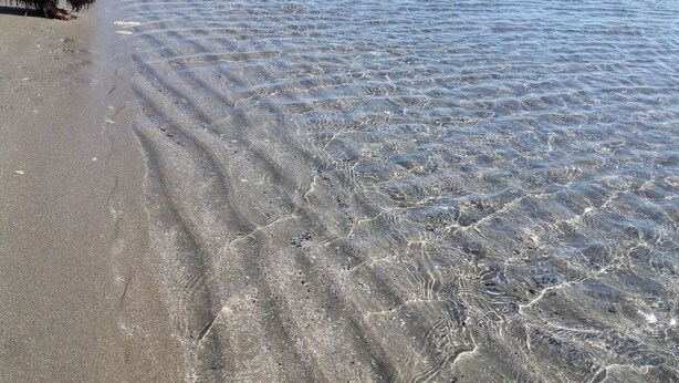 En strand med klart, lavt vand og synlige sandmønstre.