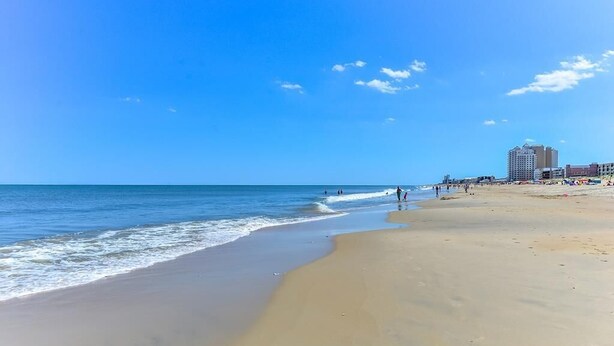 A beach with a clear sky, a sandy shore, and a few buildings in the background.