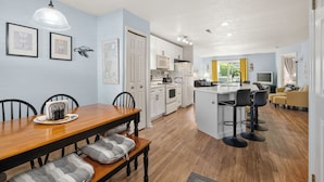 A modern kitchen with white cabinets, a dining area with a wooden table and chairs, and a living area with a sofa and a TV.