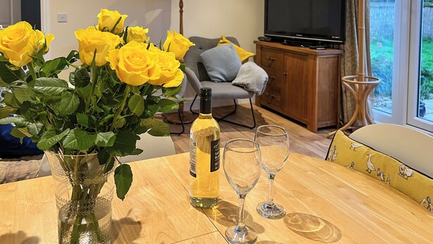 A living room with a wooden table, a vase of yellow roses, a bottle of wine, and two wine glasses.