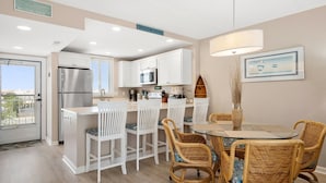 Une cuisine moderne avec des meubles blancs, un réfrigérateur en acier inoxydable et un coin repas comprenant des chaises en osier et une table en verre.