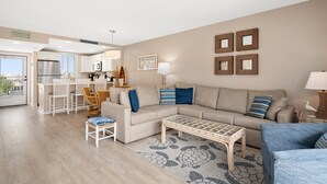 Un salon moderne comprenant un canapé, une table basse et des fauteuils, un coin cuisine avec un réfrigérateur et un ensemble de salle à manger, ainsi qu’un balcon avec vue.