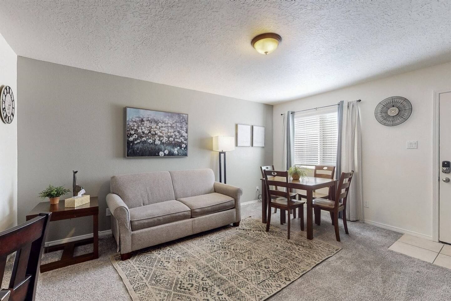 Una sala de estar con un sofá color beige, una mesa de comedor con sillas, un tapete estampado y un cuadro en la pared.
