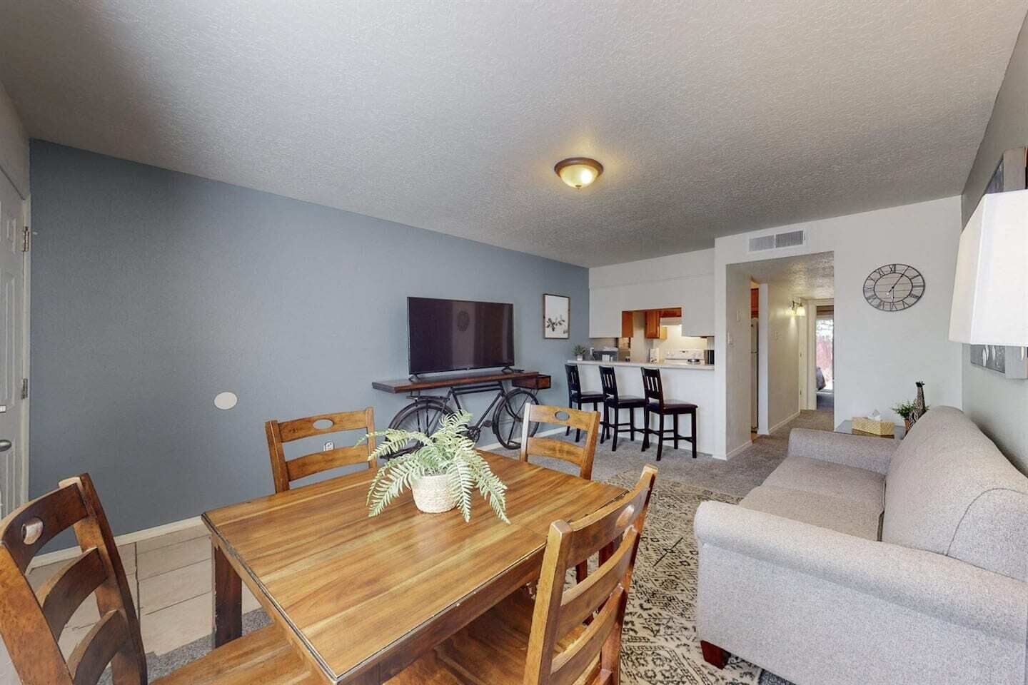 Un coin repas avec une table et des chaises en bois, un téléviseur et, au fond, une cuisine.