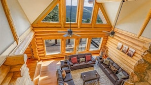 Ein Holzblockhaus-Interieur mit großem Wohnbereich, ausgestattet mit einer Sofagarnitur, einem Couchtisch und einem Kamin. Mehrere Fenster bieten Ausblick auf die umliegende Landschaft.