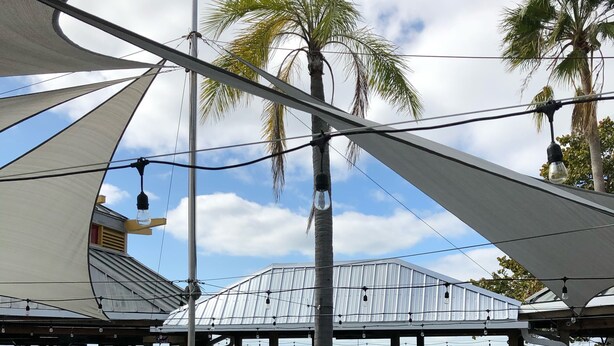 An outdoor seating area with tables and chairs, a covered section with a view of the sea, and a building with a metal roof in the background.