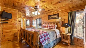 Een slaapkamer in een houten chalet met een bed, een plafondventilator, een televisie en een raam met gordijnen.