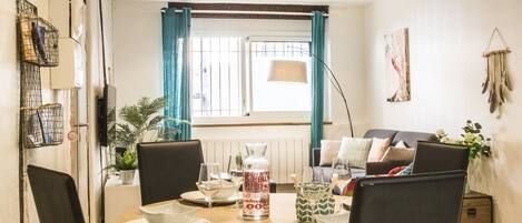 A dining area with a round table set for four, a sofa in the background, and a window with curtains.