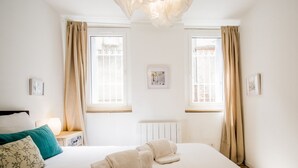 Un dormitorio con una cama grande, dos ventanas con cortinas, un radiador y un cuadro colgado en la pared.
