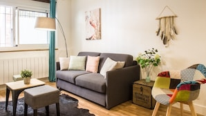 Una sala de estar con un sofá gris, una mesita, un sillón colorido, una lámpara de pie y una obra de arte en la pared.