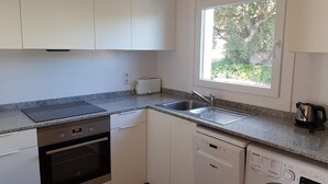 Ett kök med vita skåp, en inbyggd ugn, en diskmaskin, en brödrost och ett fönster med utsikt över grönområden.