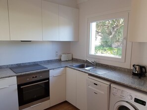 A kitchen with white cabinets, a built-in oven, a dishwasher, a toaster, and a window with a view of greenery. - K-4 Apartment in a quiet area, with swimming pool (Calella de Palafrugell)