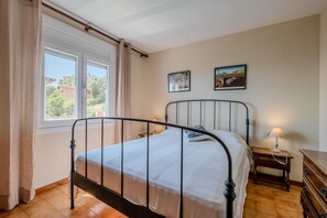 A bedroom with a metal-framed bed, a wooden nightstand with a lamp, a dresser, and two framed artworks on the wall. - K-4 Apartment in a quiet area, with swimming pool (Calella de Palafrugell)