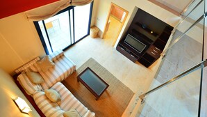 A modern living room with a sofa, a coffee table, and a TV. There is a large window and a door leading to another room.