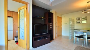 A modern living room with a large flat-screen TV, built-in shelving, and a dining area with a glass table and chairs.