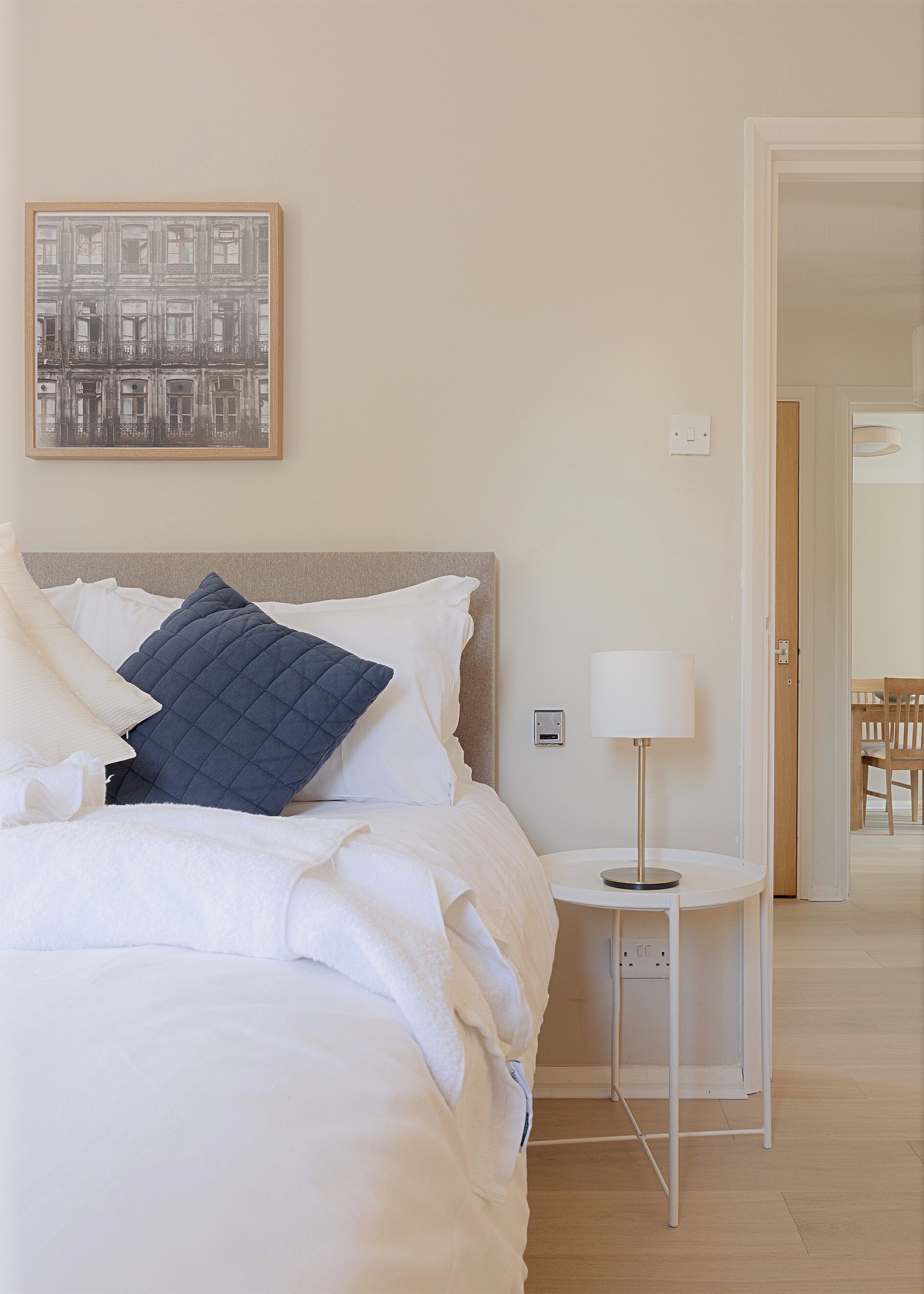 A neatly made bed with white and blue bedding, a bedside table with a lamp, and a framed picture on the wall.