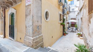 Un estrecho callejón con un edificio amarillo, un pequeño espacio al aire libre con mesas y sillas, y un letrero sobre una puerta.