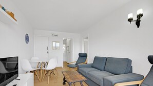 A modern living room with a grey sofa, a wooden coffee table, white chairs, a TV, and a wall-mounted light fixture.