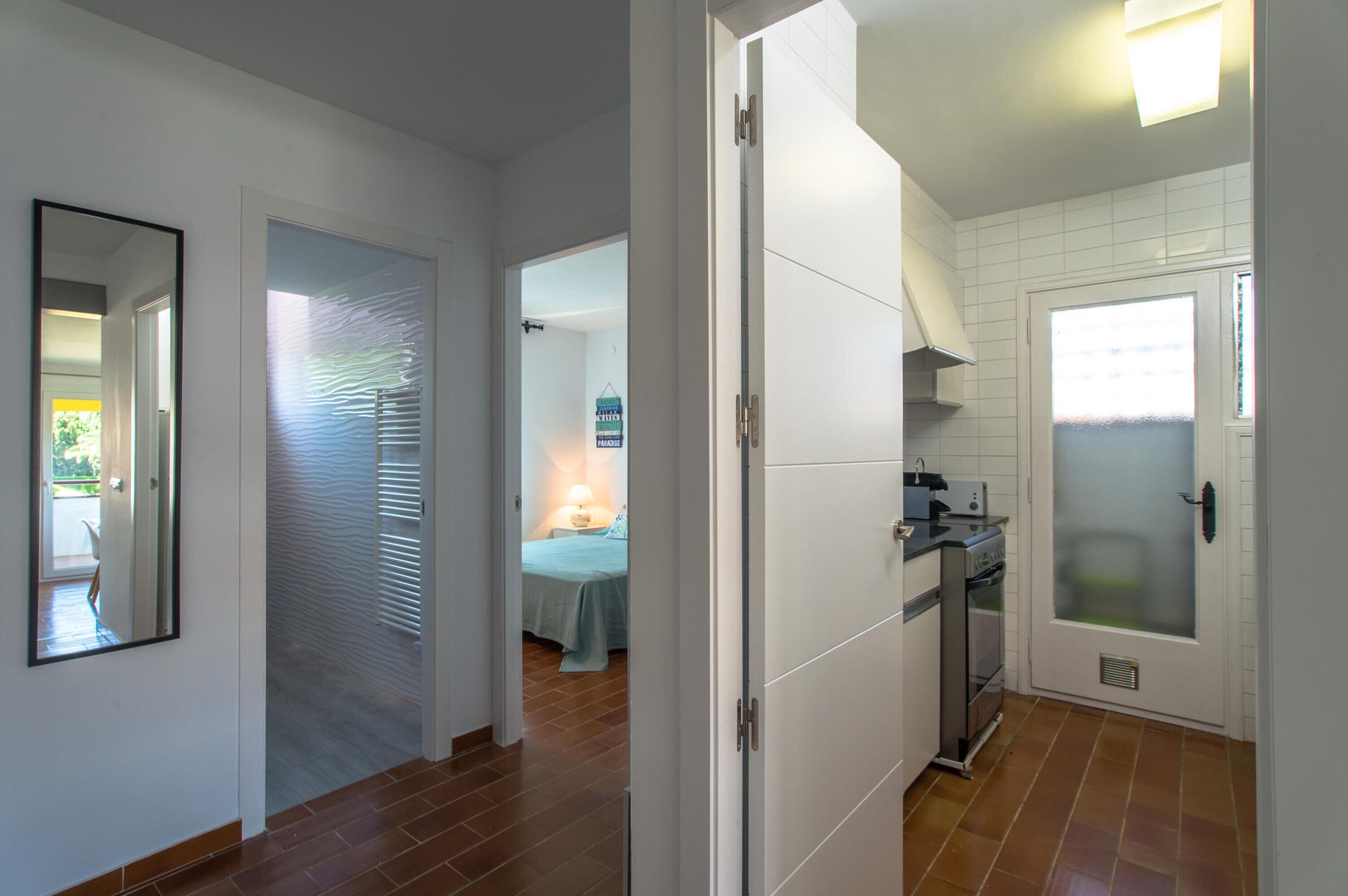 A hallway leading to a bedroom with a bed and a nightstand, a kitchen with a stove and oven, and a bathroom with a frosted glass door.