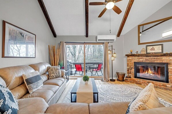 A cozy living room with a leather sofa, a fireplace, and a large window overlooking a deck.