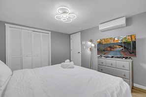 A bedroom with a bed, a white dresser, a TV on the wall, and a ceiling light fixture.