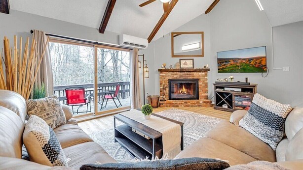 A spacious living room with a fireplace, a ceiling fan, and a large window overlooking a deck.