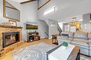 Cozy living room with a fireplace, comfortable sofa, and dining area, featuring a large window and staircase.