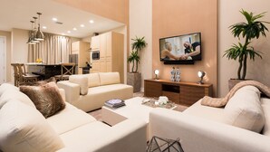 Un salon moderne avec un canapé, une table basse et un téléviseur. On aperçoit au fond une cuisine avec des meubles en bois et un coin repas.
